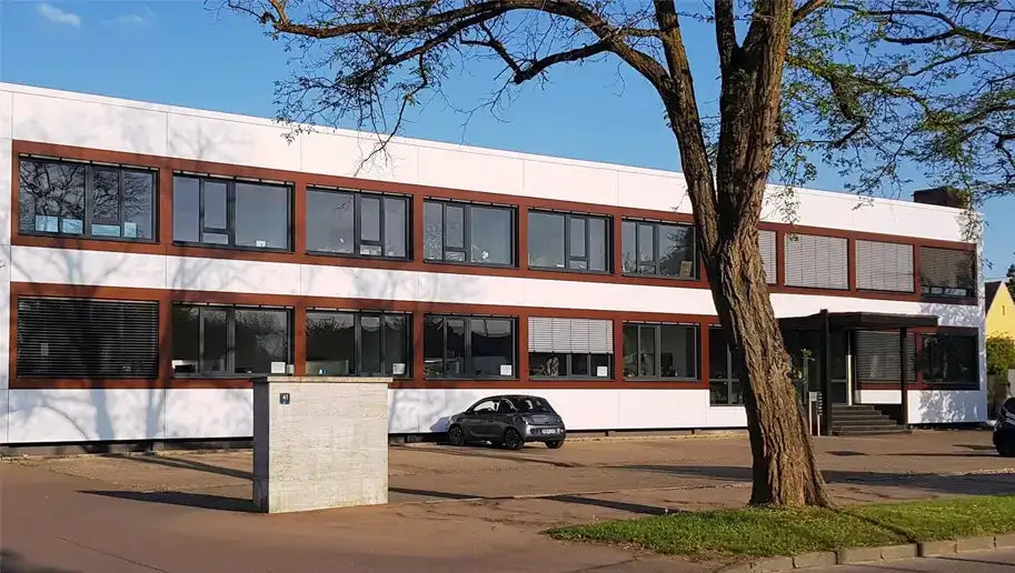 Modernes Bürogebäude mit großem Baum im Vordergrund und mehreren geparkten Autos auf dem Parkplatz davor.